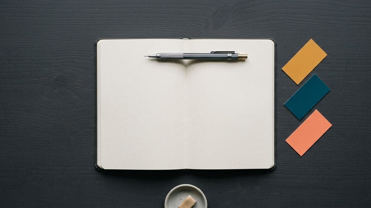 An open notebook with completely blank pages on a dark wooden desk, a mechanical pencil resting across it, and three small colour swatches in amber, teal and coral arranged in a diagonal row beside it.