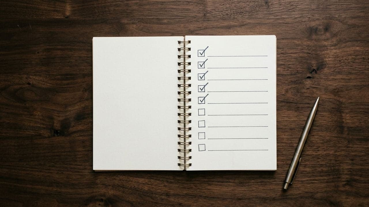 An open spiral-bound notebook on a dark wooden desk, photographed from above. A column of nine small checkboxes runs down the page; five carry a hand-drawn tick and four are empty. The ruled lines beside them are blank. A slim pen rests on the desk alongside.